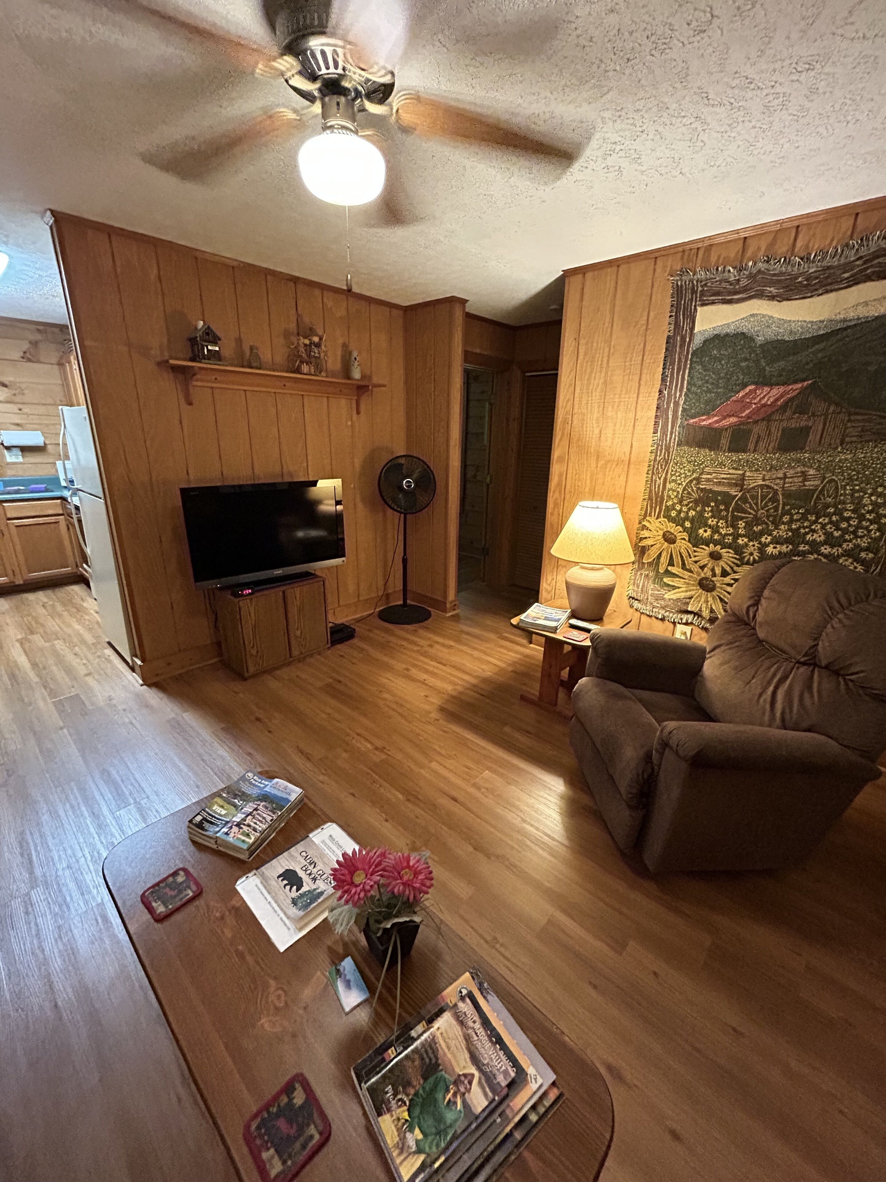 Una sala de estar con paredes de madera, un ventilador de techo, un televisor de pantalla plana, una mesa de centro con revistas y un florero, y un sofá.