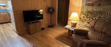 Una sala de estar con paredes de madera, un ventilador de techo, un televisor de pantalla plana, una mesa de centro con revistas y un florero, y un sofá.