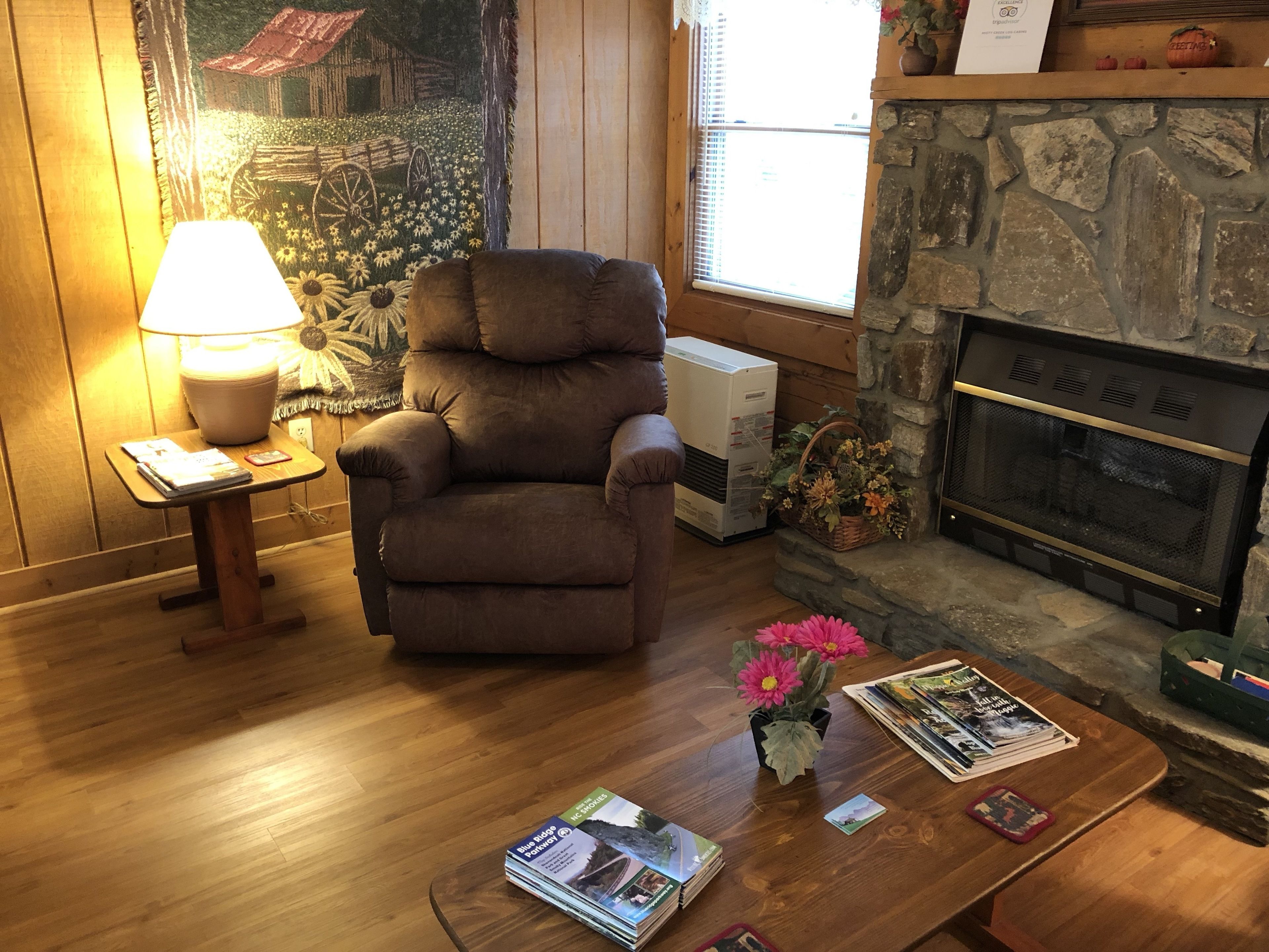 A cozy living room with a fireplace, a comfortable armchair, a table with magazines, and a lamp.