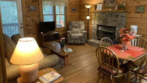 A cozy cabin interior with a fireplace, wooden walls, a television, a comfortable armchair, a dining table with chairs, and a lamp. - Misty Creek Log Cabins (Maggie Valley)