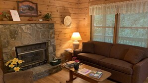 A cozy cabin interior with a stone fireplace, wooden walls, a sofa, a coffee table with books, and a window with blinds. - Misty Creek Log Cabins (Maggie Valley)