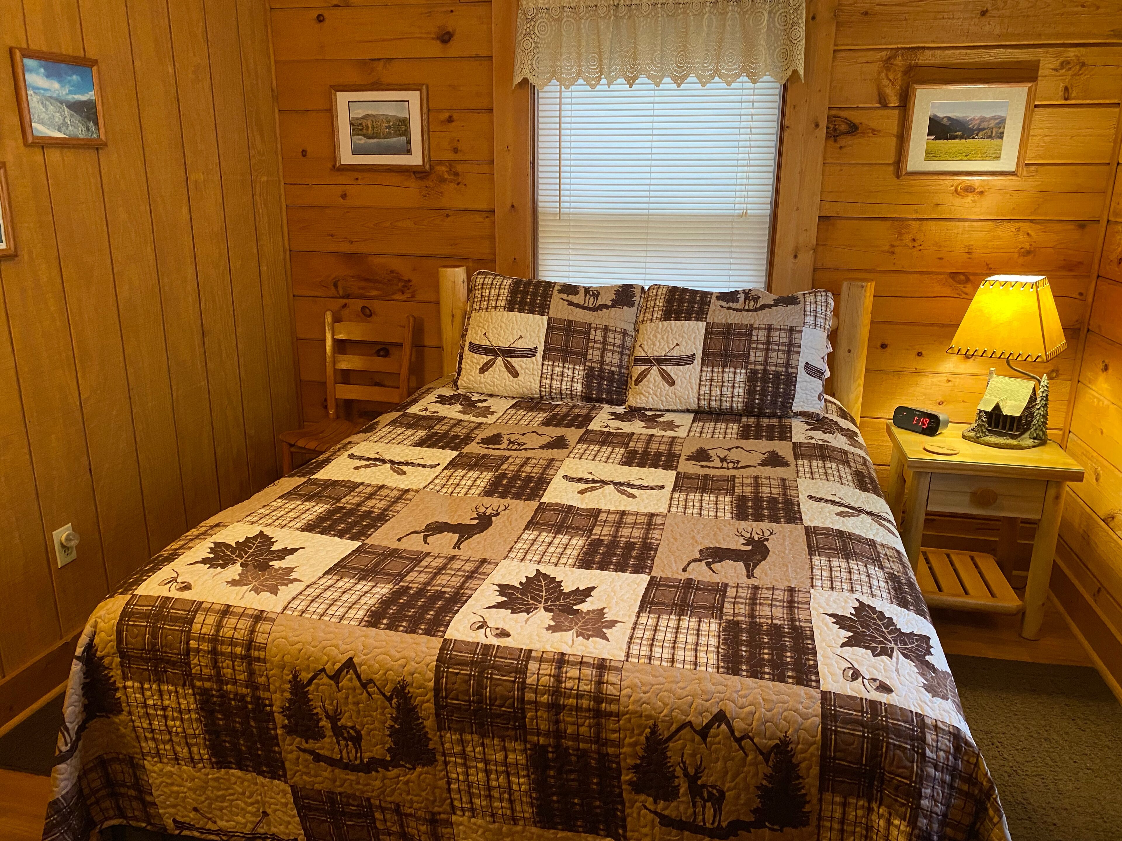 Ein gemütliches Schlafzimmer im Holzstil, mit einem Bett mit einem Quilt-Muster von Hirschen und Blättern, einem Nachttisch mit Lampe und gerahmten Bildern an den Wänden.
