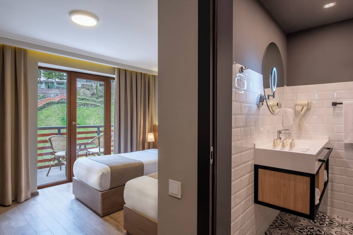 Habitación de hotel con balcón, cama doble, baño con lavamanos y espejo modernos, y vistas a un paisaje verde.