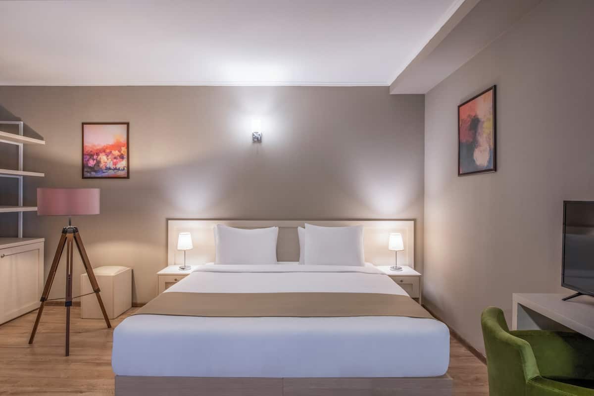 Habitación de hotel con una cama grande, lámparas de noche, un escritorio con televisor, una silla verde y obras de arte en las paredes.