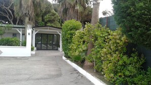 A building entrance with a covered walkway, surrounded by lush greenery and palm trees. - Hotel LePinède (Giugliano in Campania)