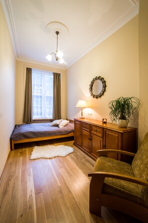Une chambre à coucher comprenant un lit, une commode, une chaise, une lampe, un miroir et une fenêtre avec des rideaux.