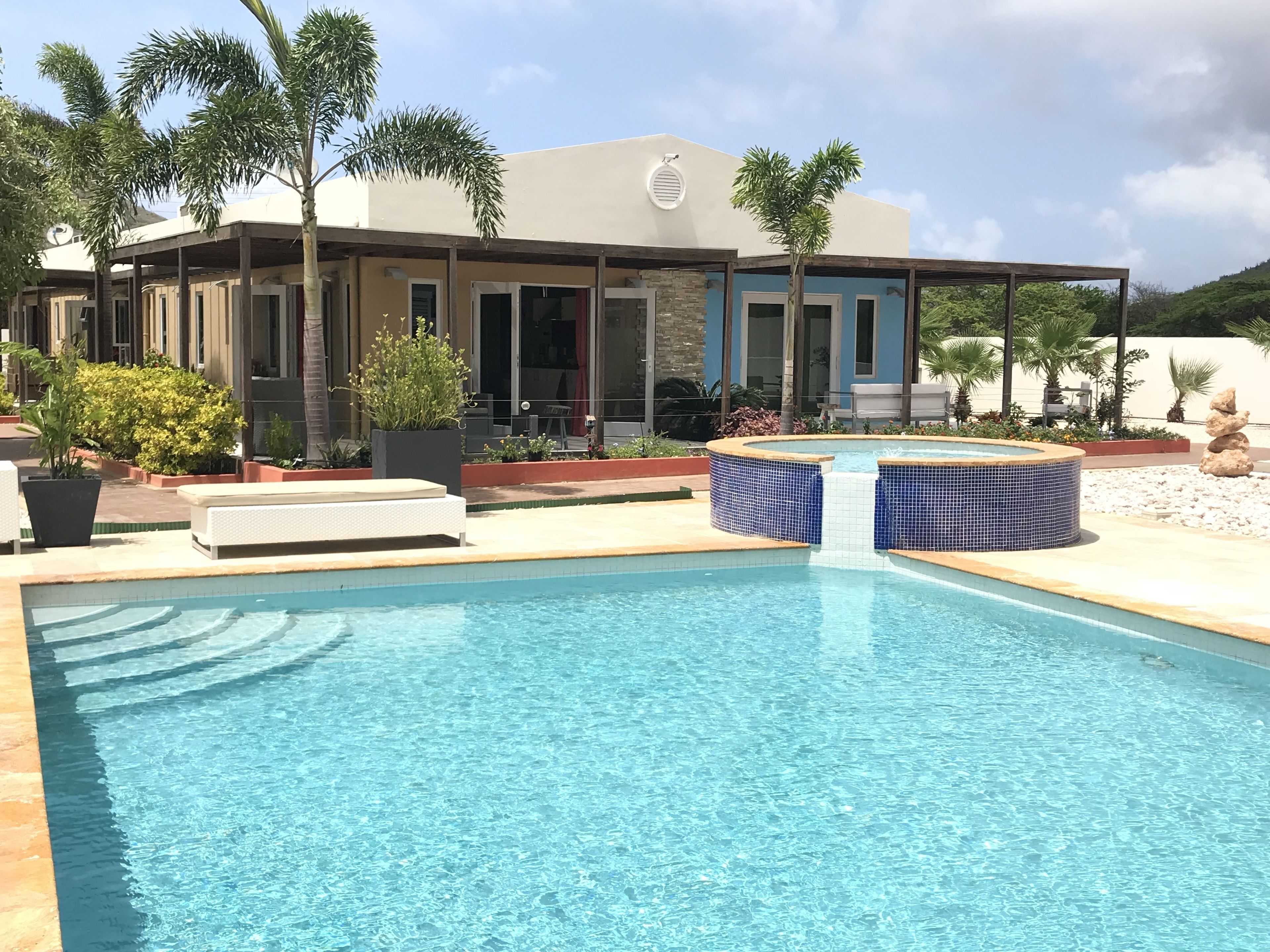 Un área de piscina moderna con una piscina azul cristalina, rodeada por un patio con mobiliario y plantas en macetas.