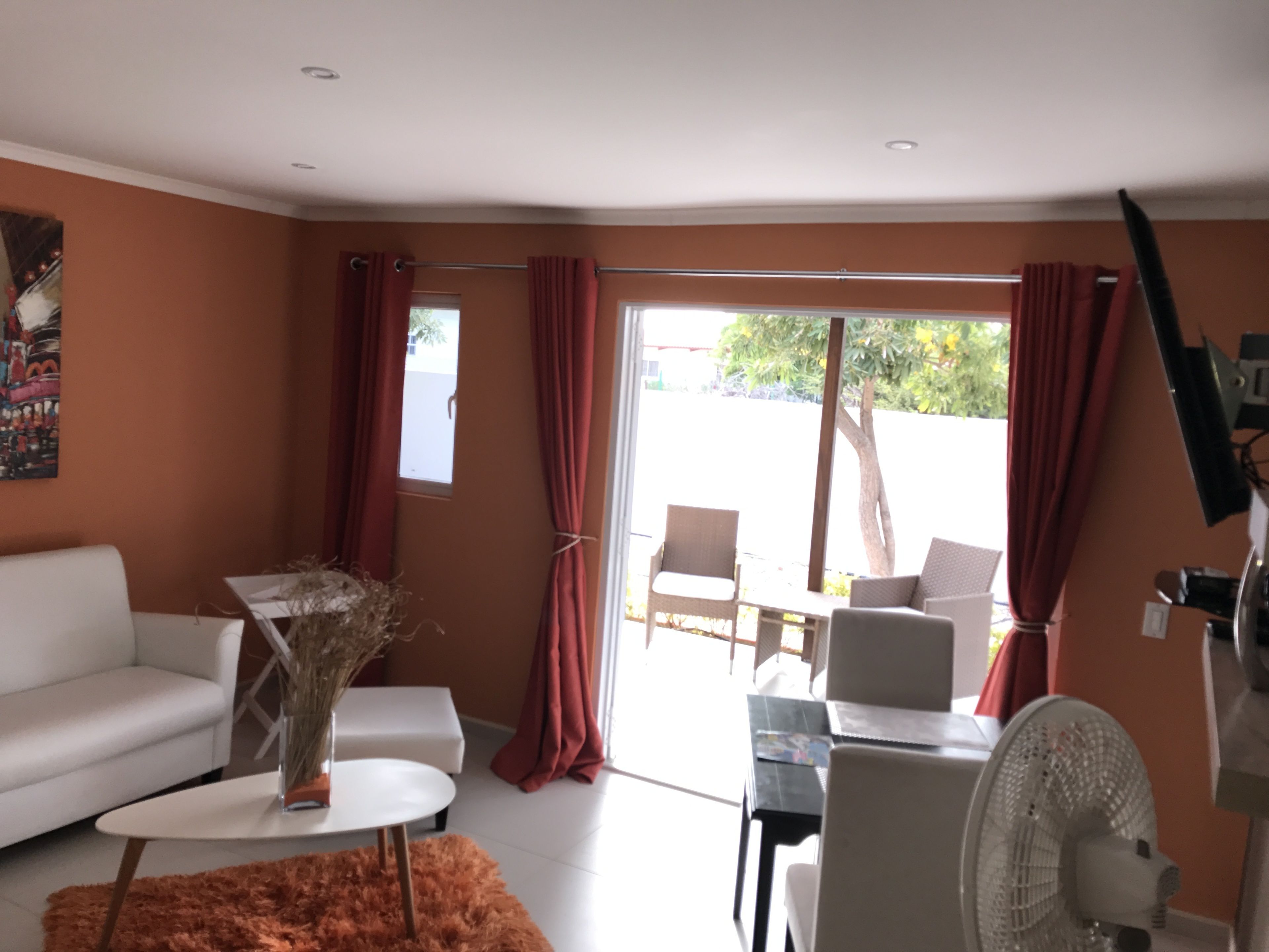 Una sala moderna con un sofá blanco, una mesita redonda y un ventilador. Se ve una ventana con cortinas rojas y vistas al exterior.