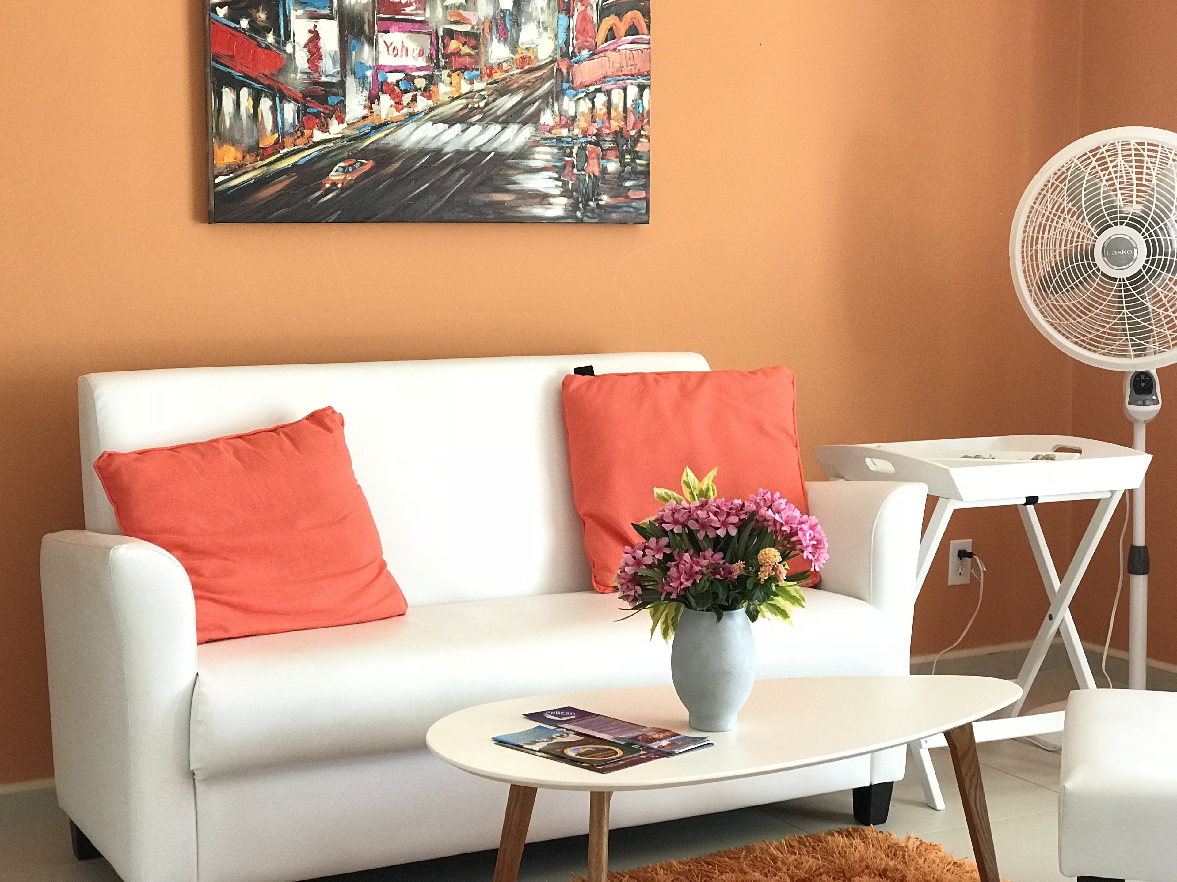 A modern living room with a white sofa, red cushions, a small round table with a vase of flowers, and a standing fan.