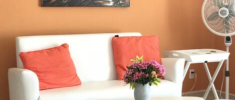 A modern living room with a white sofa, red cushions, a small round table with a vase of flowers, and a standing fan.
