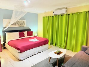 A neatly made bed with red and white bedding, a bedside table with a green apple and a wooden tray, and a bright green curtain. - Malika Apartments (Sabana Westpunt)