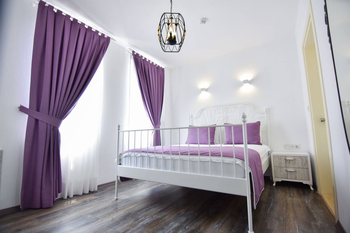 A bedroom with a white metal bed frame, purple bedding, a white nightstand, and a hanging light fixture.