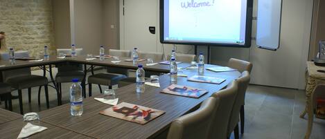 Sala de conferências com uma tela grande exibindo "Welcome!" e um quadro branco. Mesas com garrafas de água e papéis.