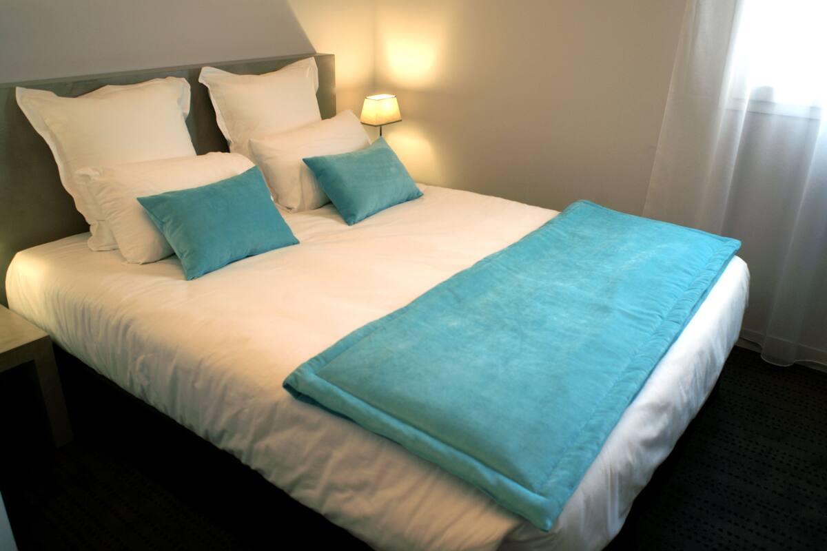 Een keurig opgemaakt bed met wit en blauw beddengoed, een bedlampje en een raam met gordijnen.