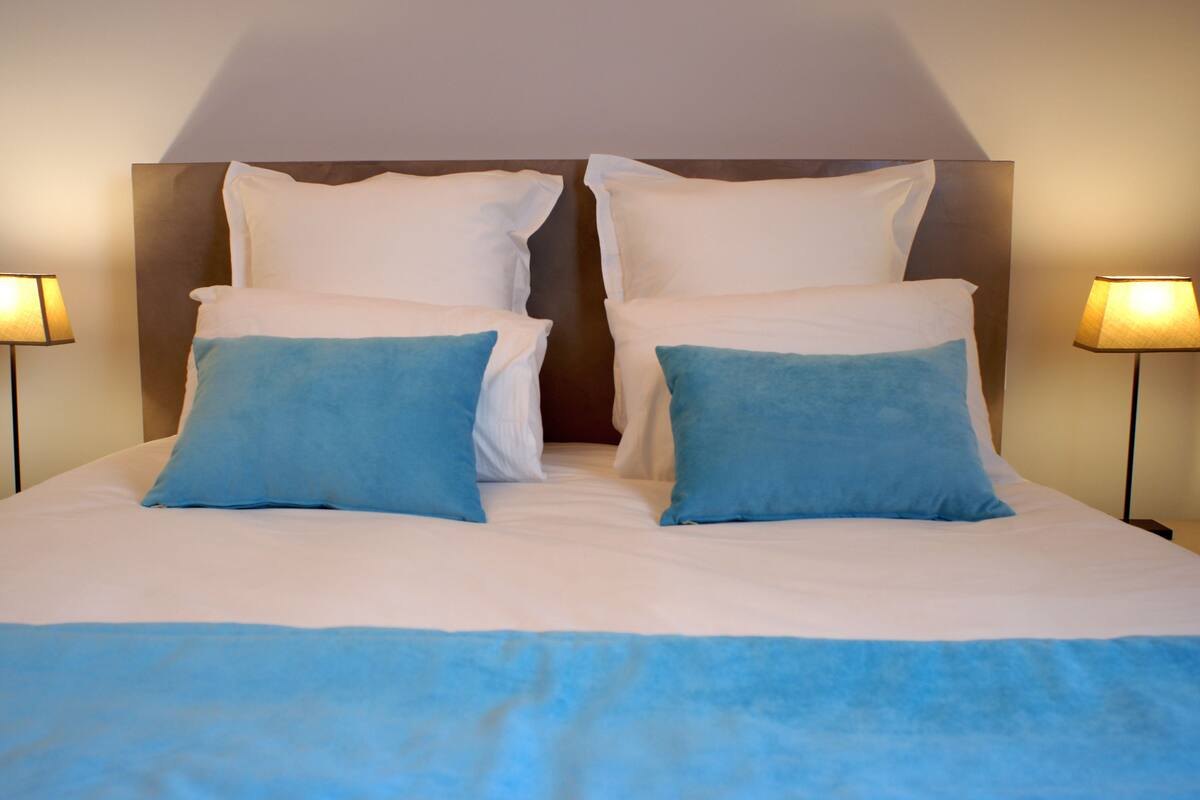 Een netjes opgemaakt bed met wit- en blauwe beddengoed, twee lampen aan weerszijden, en een houten hoofdbord.