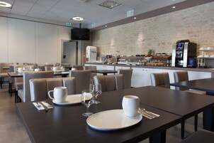 Un comedor moderno con mesas dispuestas para dos, con platos, tazas y cubiertos blancos.