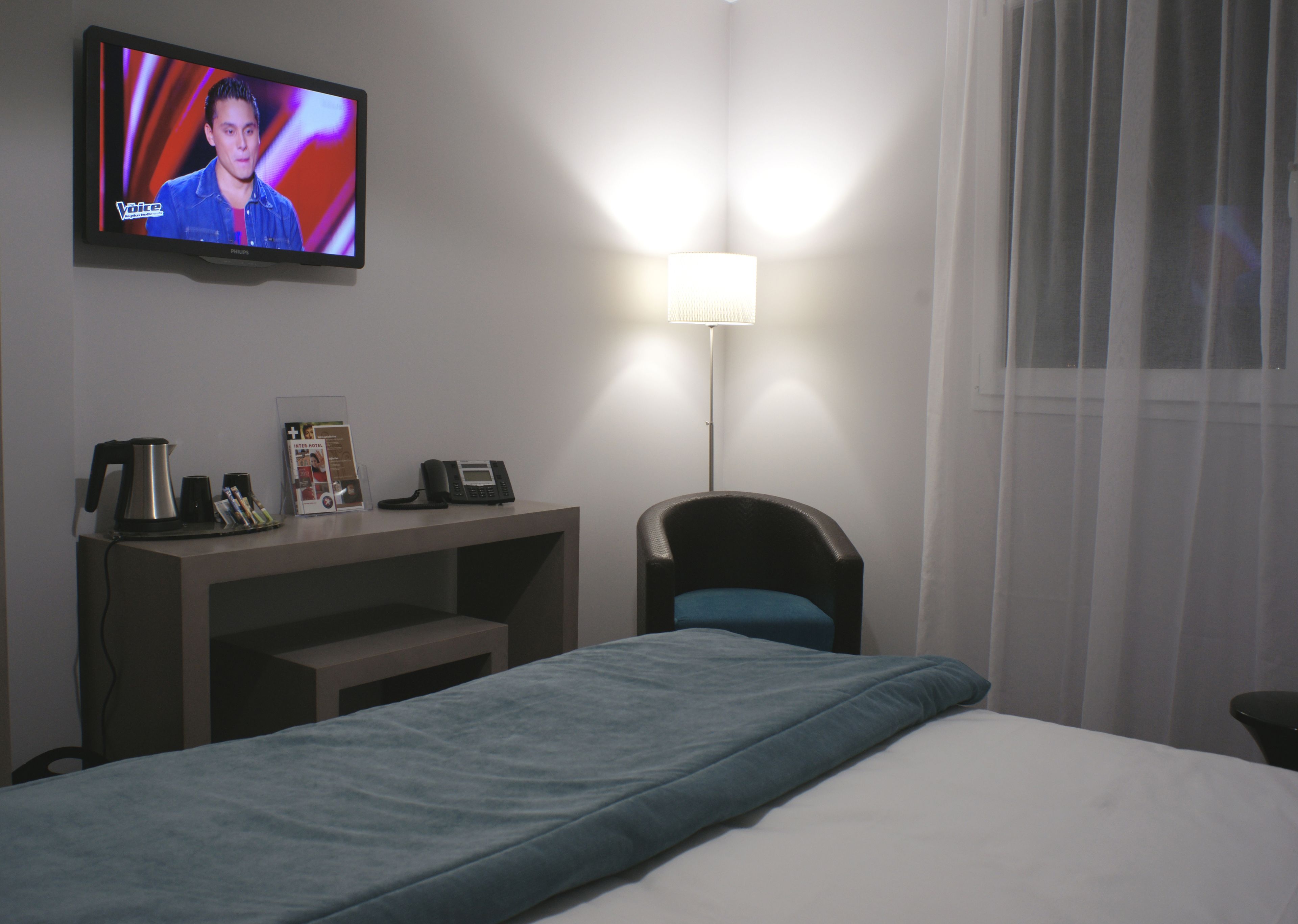 Habitación de hotel con cama, escritorio, teléfono, cafetera y televisor mostrando un programa.