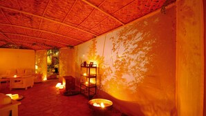 A room with a thatched roof, stone floor, and a fire pit. There are sofas and a shelf with candles. - Lycia Hotel (Kas)