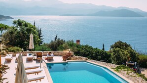 A swimming pool with lounge chairs and umbrellas, surrounded by greenery and a view of the sea and mountains. - Lycia Hotel (Kas)