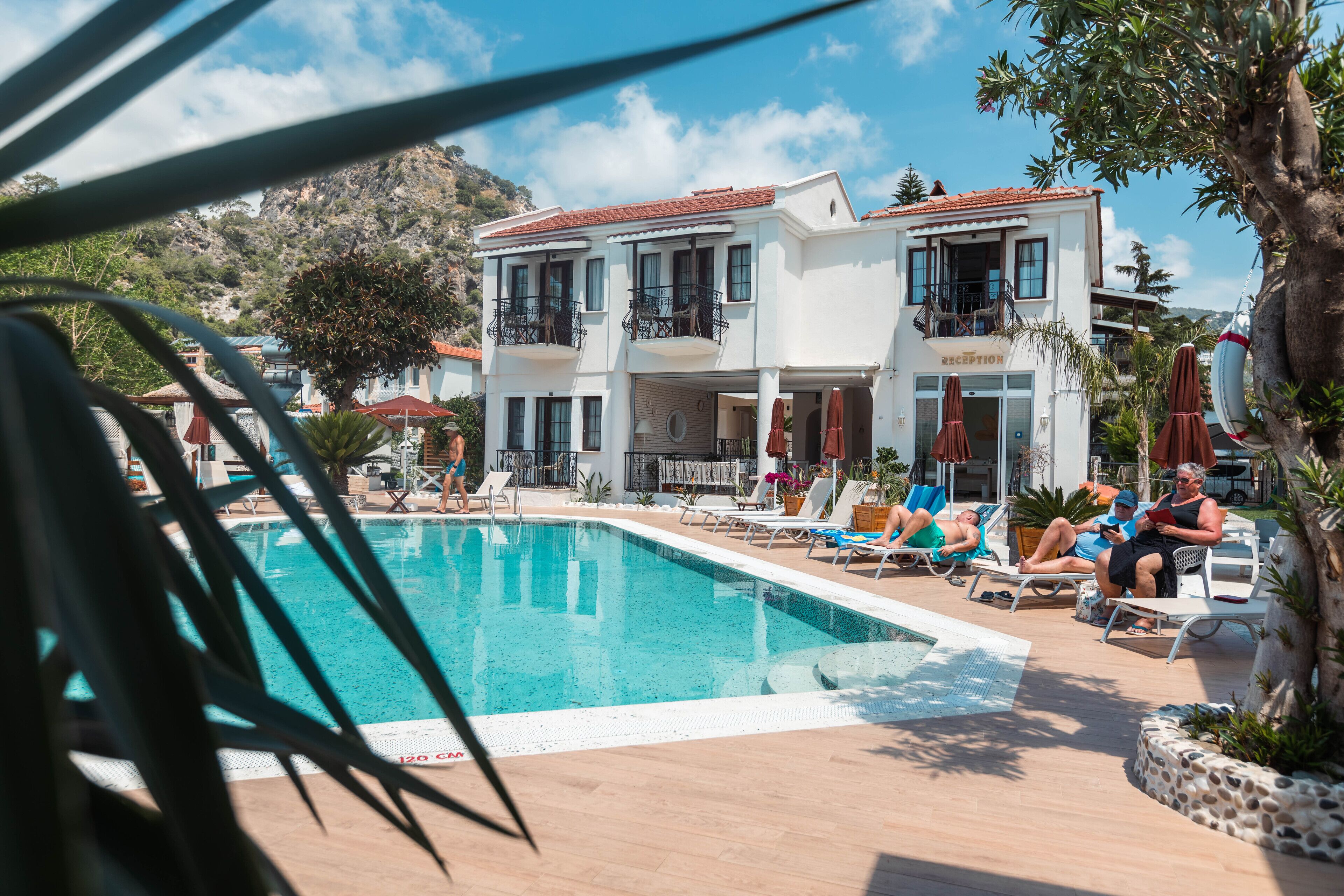 A poolside area with lounge chairs and umbrellas, a reception building, and a mountainous backdrop.