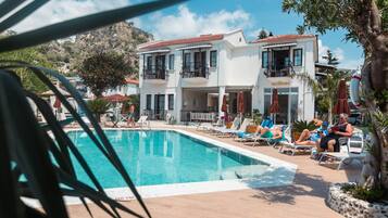 A poolside area with lounge chairs and umbrellas, a reception building, and a mountainous backdrop.