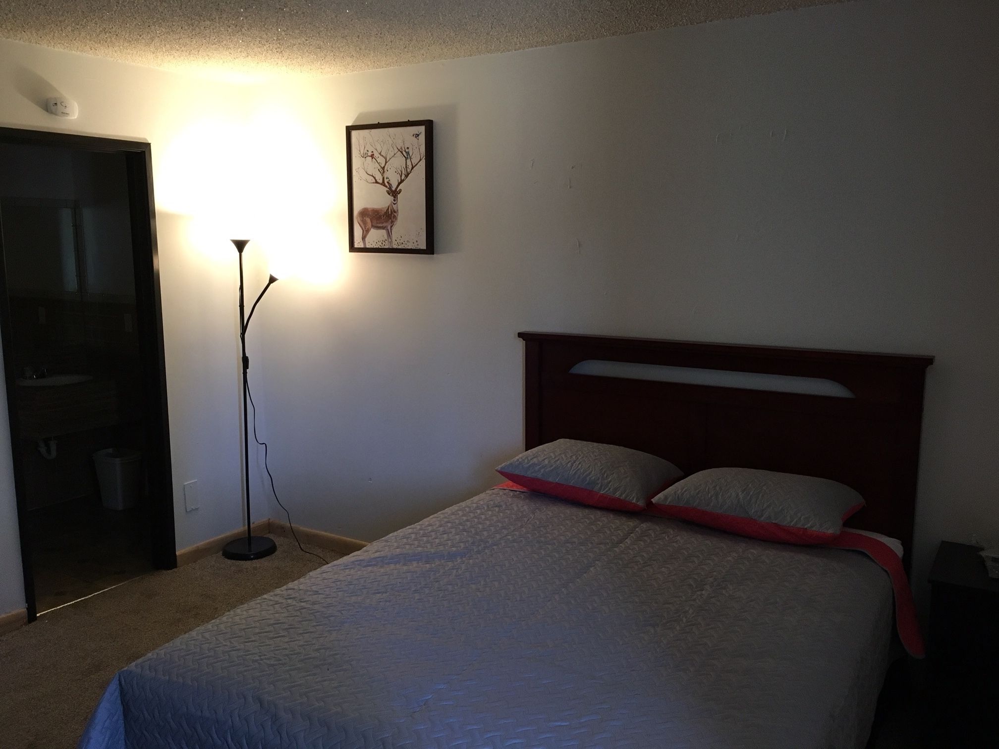 A bedroom with a bed, a nightstand, a lamp, a framed picture on the wall, and a door to a bathroom.