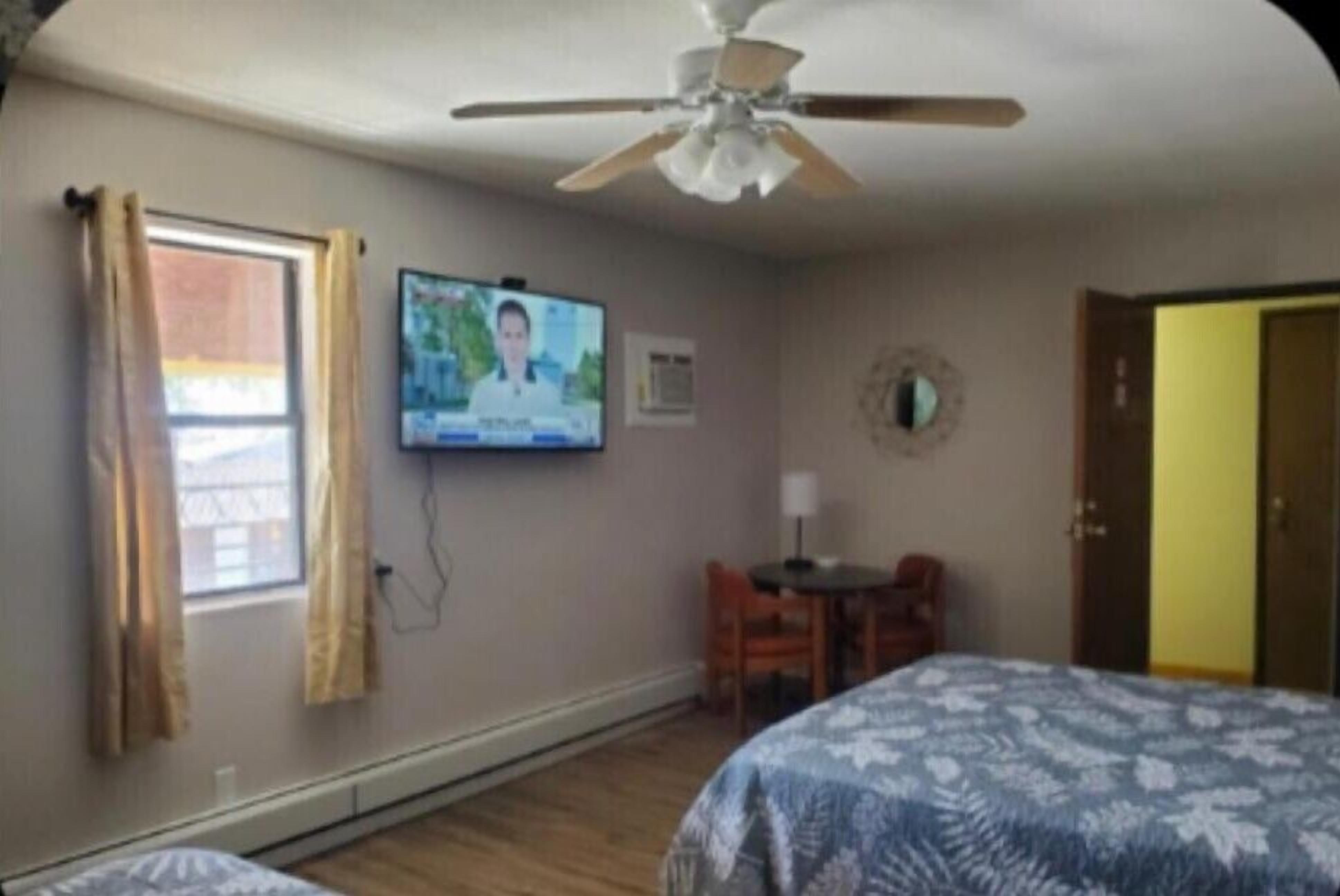 Une chambre d’hôtel avec deux lits, un ventilateur de plafond, une télévision, une petite table avec une chaise et une fenêtre avec des rideaux.