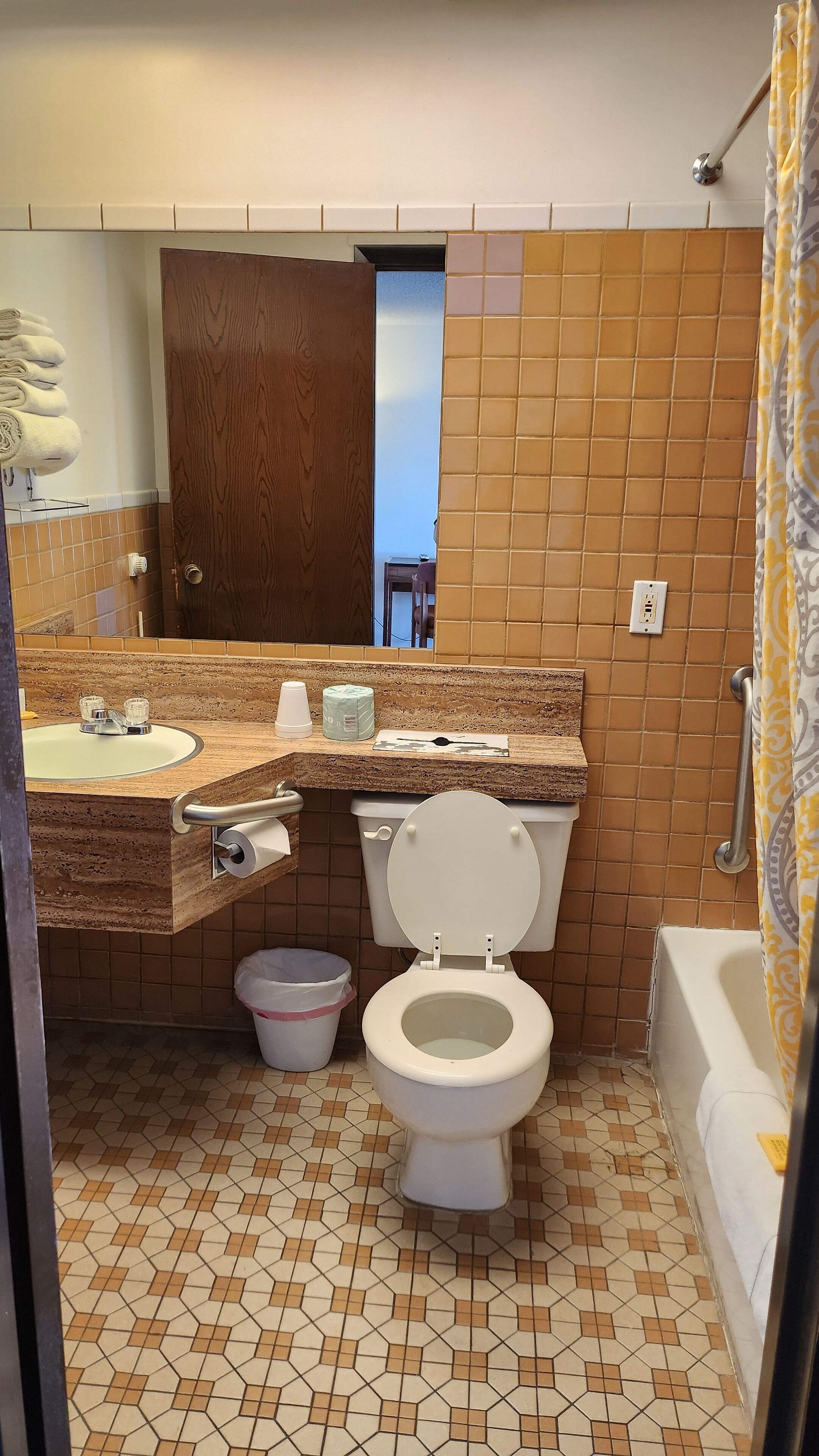 Casa de banho com sanita, lavatório e banheira. Tem uma porta de madeira, um porta-toalhas e um cesto de lixo.