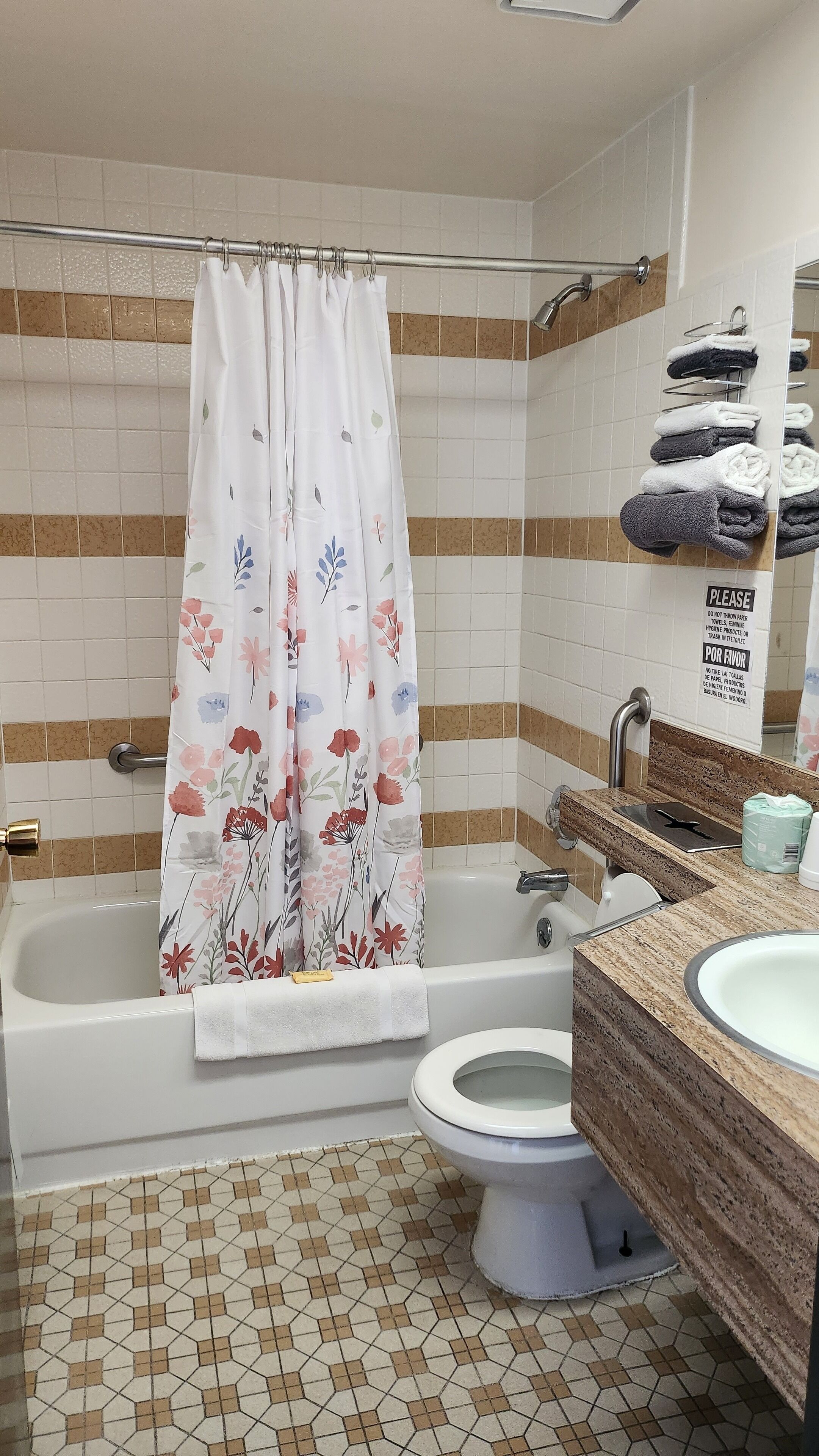 Une salle de bain avec une baignoire, des toilettes et un lavabo. Il y a un rideau de douche orné de motifs floraux, et des serviettes sont suspendues à un porte-serviettes.