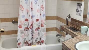 Une salle de bain avec une baignoire, des toilettes et un lavabo. Il y a un rideau de douche orné de motifs floraux, et des serviettes sont suspendues à un porte-serviettes.