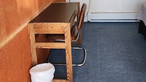A compact room with a wooden desk, a television mounted on the wall, a small window with an air conditioning unit, and a blue carpeted floor. - Golden Arrow Motel (Lamar)