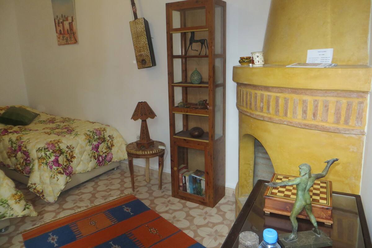A bedroom with a bed, a wooden bookshelf, a lamp, a small table with a chess piece, and a yellow wall with a fireplace.