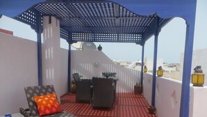 A covered outdoor seating area with a red patterned floor, a table with chairs, and potted plants. - Riad Gloria (Essaouira)