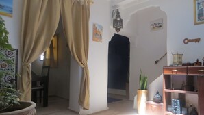 A room with a blue patterned rug, a wooden cabinet with decorative items, a potted plant, and a door with beige curtains. - Riad Gloria (Essaouira)