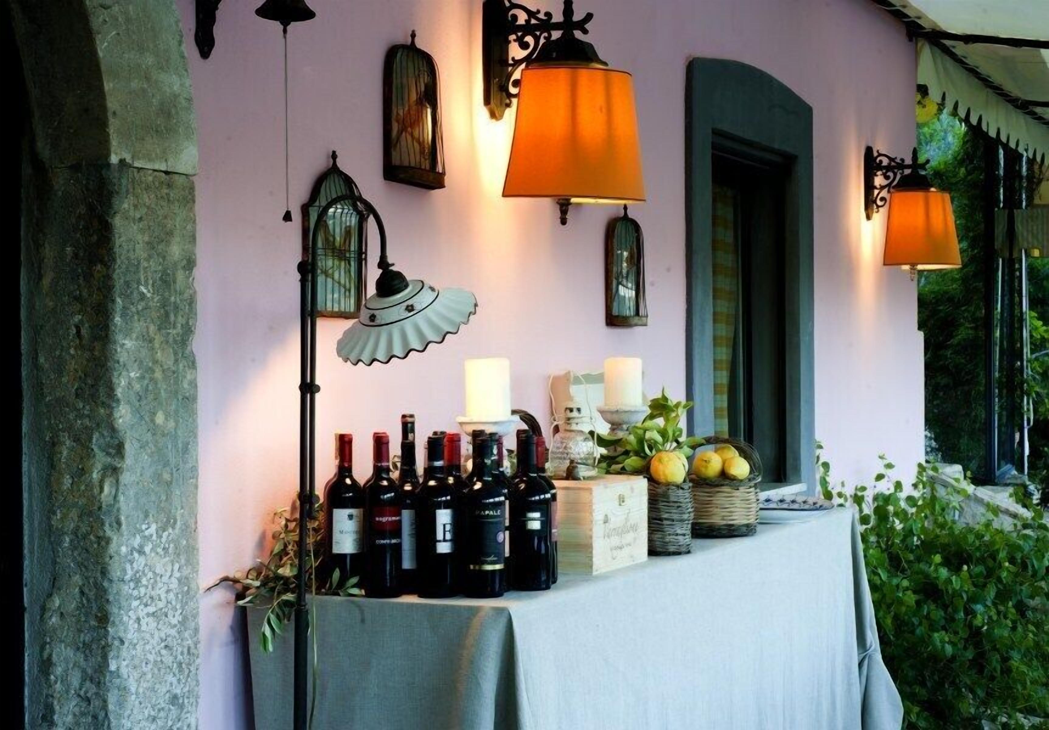 A table with wine bottles and fruits, lit by wall-mounted lamps, set against a pink wall with a green archway.