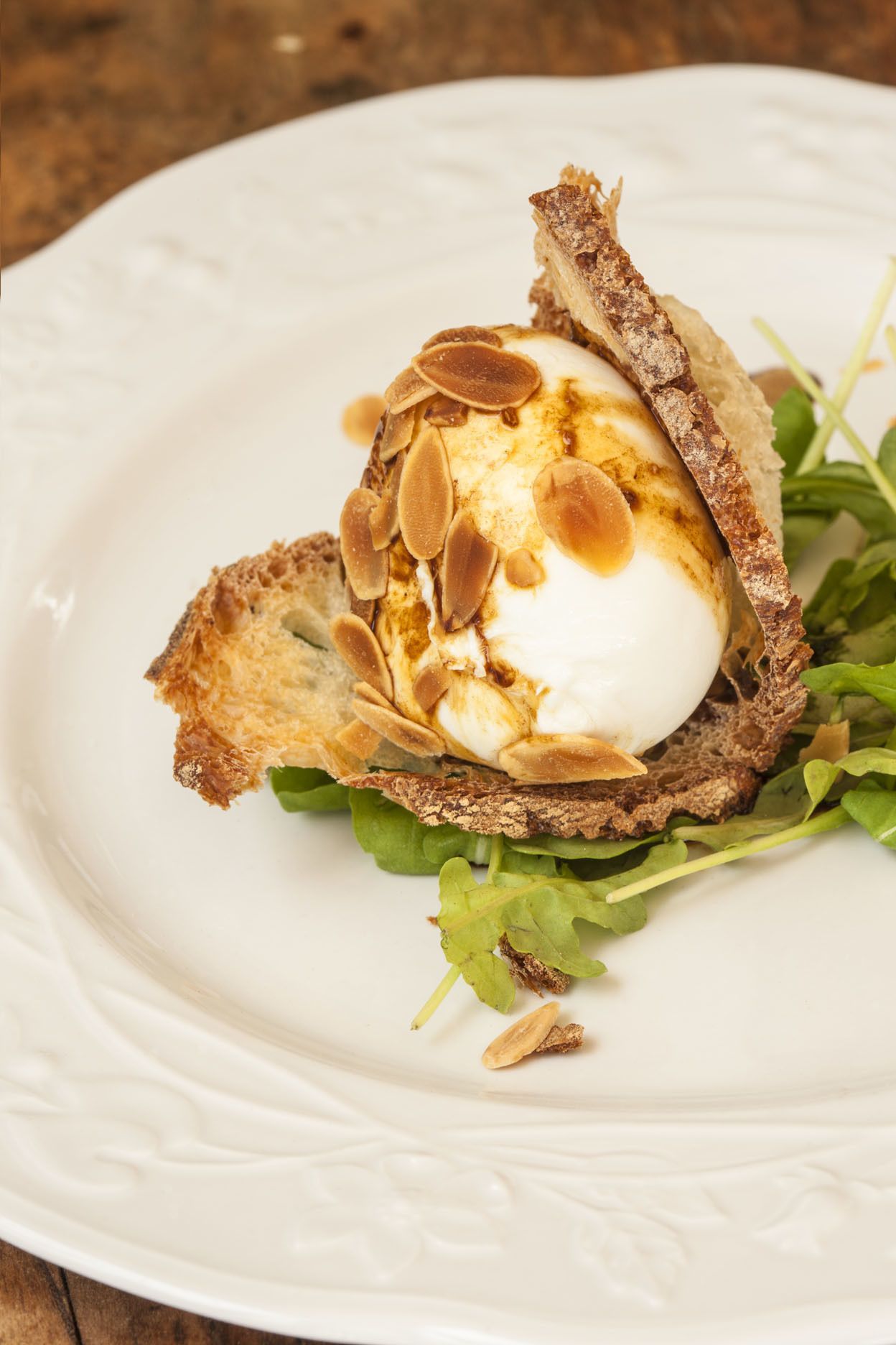A white plate with a food item consisting of a poached egg on a slice of toasted bread, topped with sliced almonds and a drizzle of sauce, accompanied by a green leafy side.