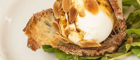 A white plate with a food item consisting of a poached egg on a slice of toasted bread, topped with sliced almonds and a drizzle of sauce, accompanied by a green leafy side.