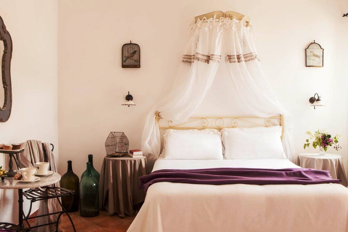 Un dormitorio con cama de dosel, una mesita con una silla, un espejo y jaulas de pájaros fijadas a la pared.