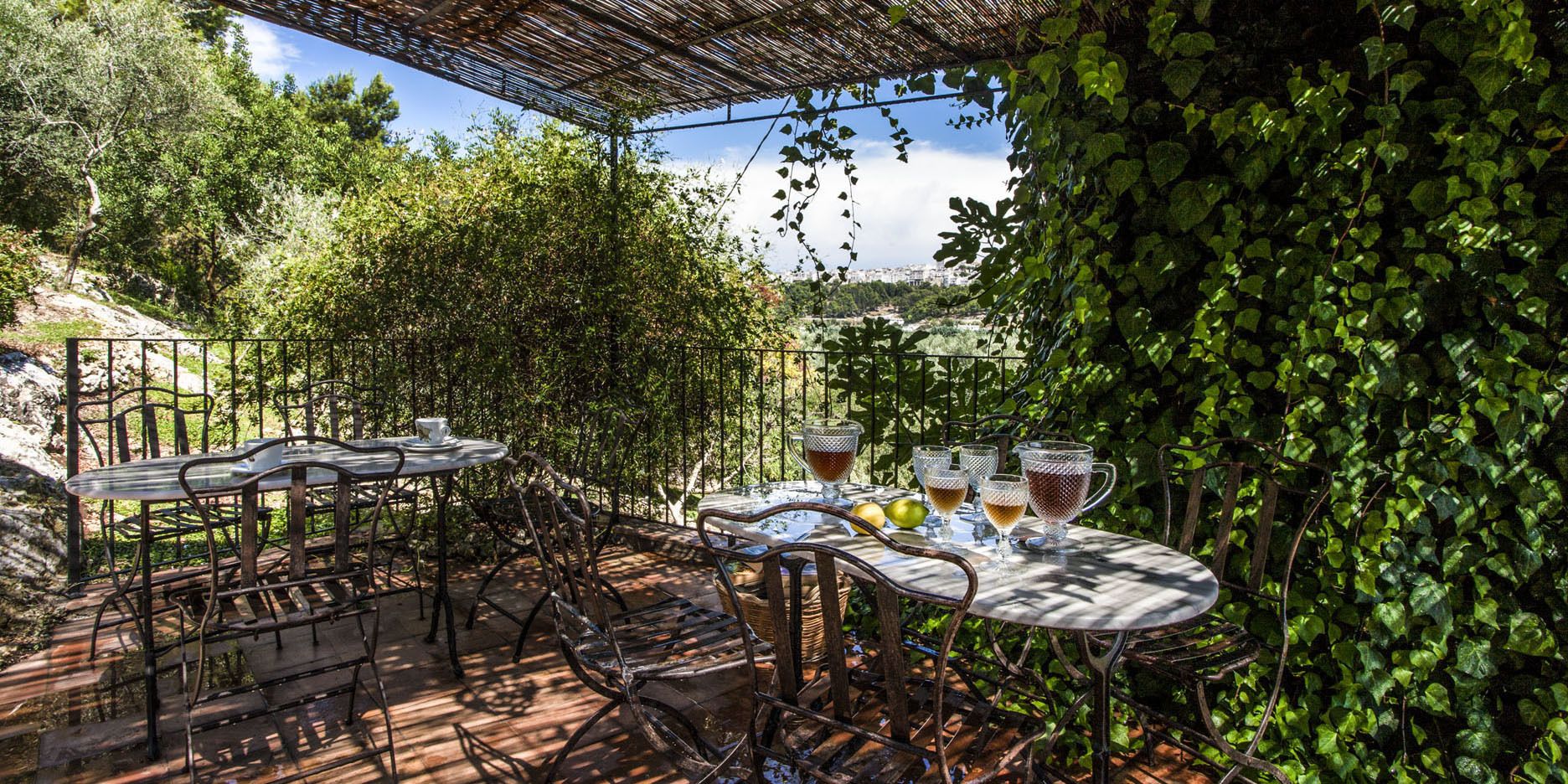 An outdoor patio with metal tables and chairs, surrounded by lush greenery and a pergola overhead.