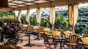 An outdoor dining area with yellow chairs and tables, large windows with curtains, and a view of a hillside.