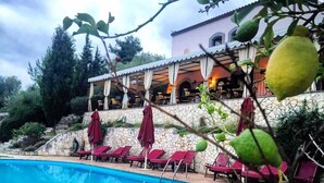 A poolside area with red lounge chairs and umbrellas, a stone wall, and a building with a tiled roof in the background.