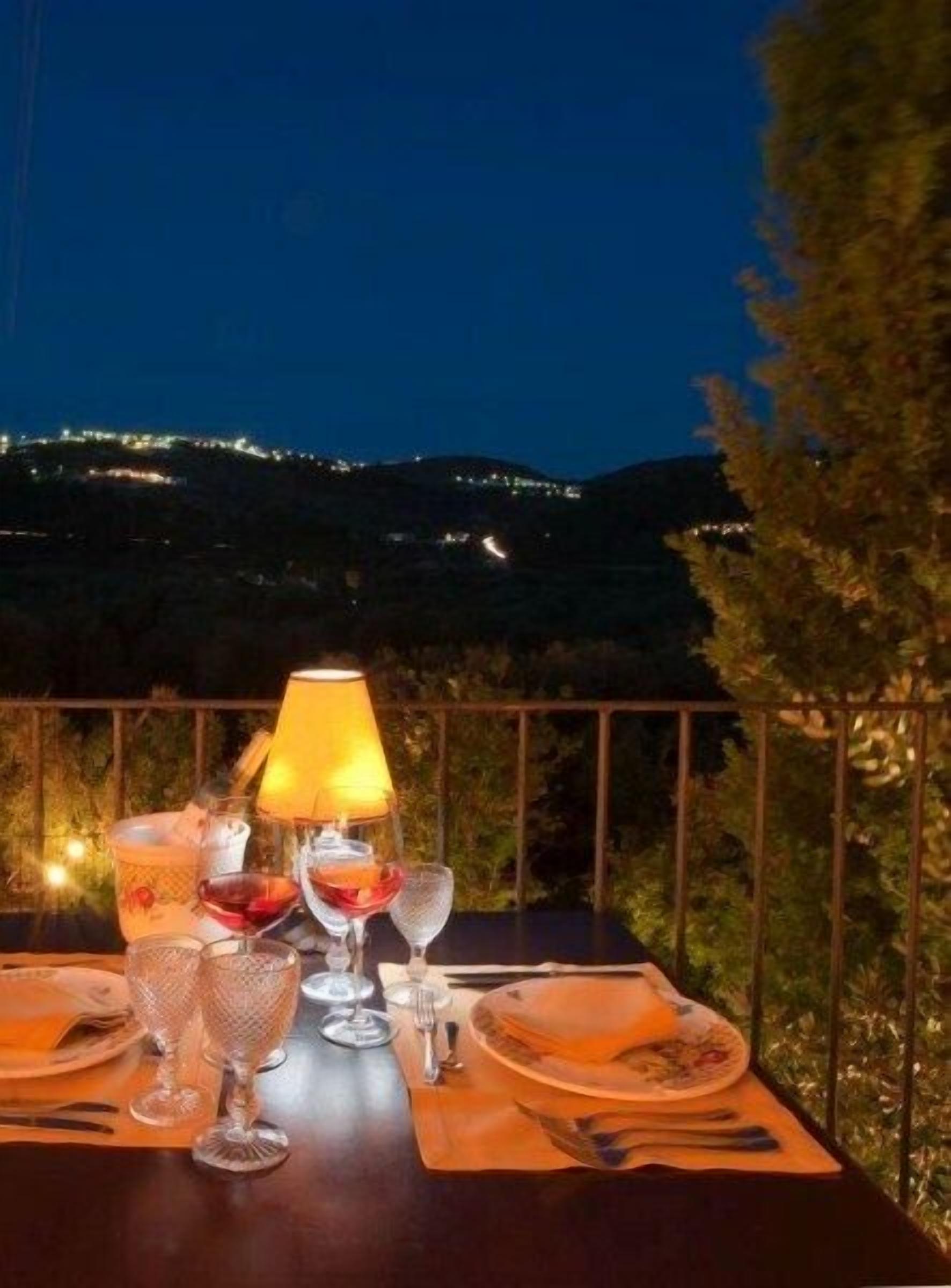 A table set for two with wine glasses, a candle, and a lamp, overlooking a mountainous landscape at night.