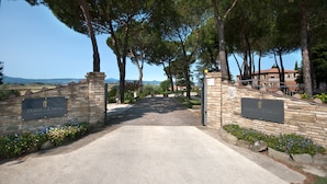 A stone entrance gate with a sign reading "Poste di Chiugi" leading to a paved path surrounded by trees and a view of the countryside. - Poste del Chiugi (Castiglione del Lago)