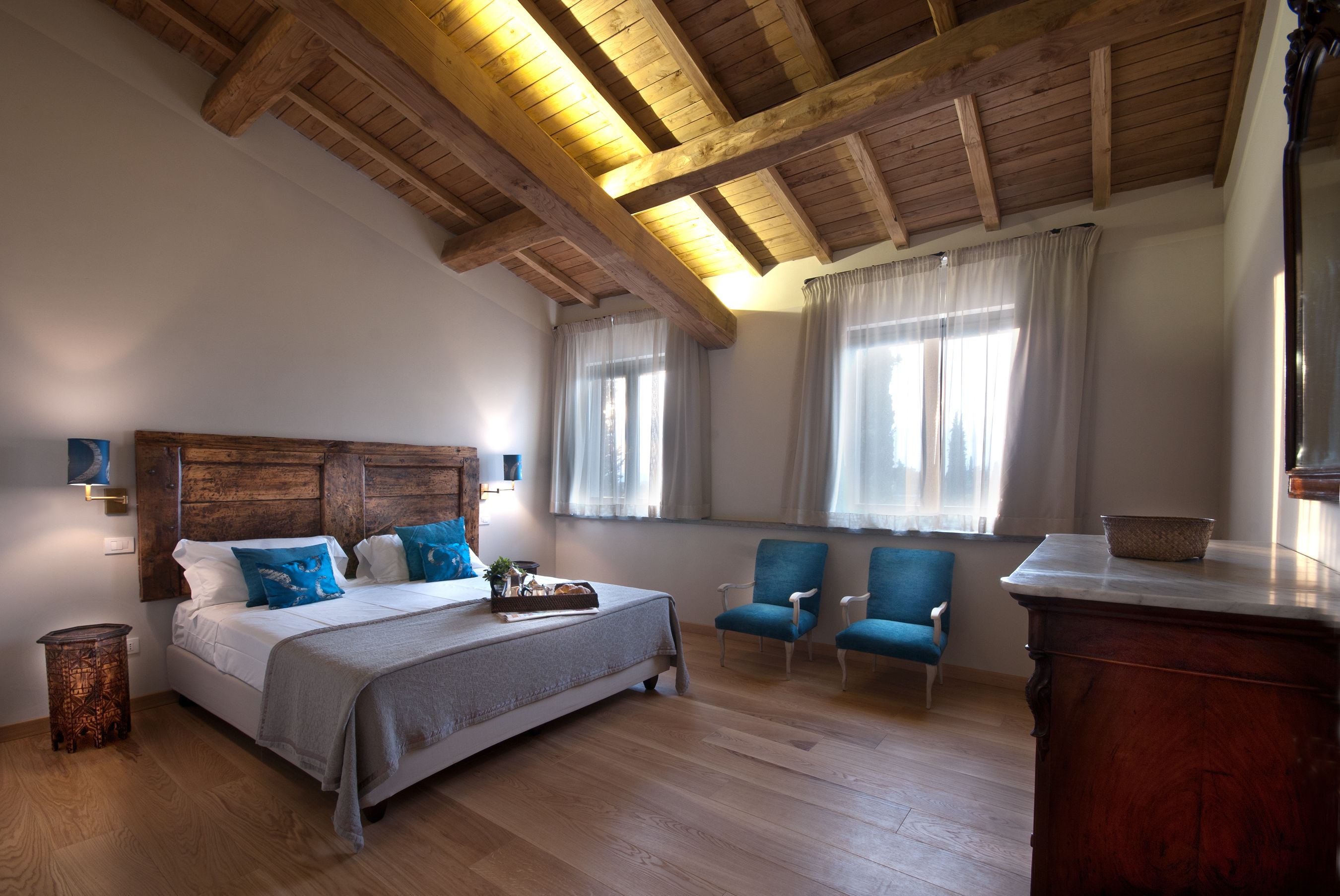 A bedroom with a wooden ceiling, a large bed with blue pillows, two blue armchairs, and a wooden nightstand.