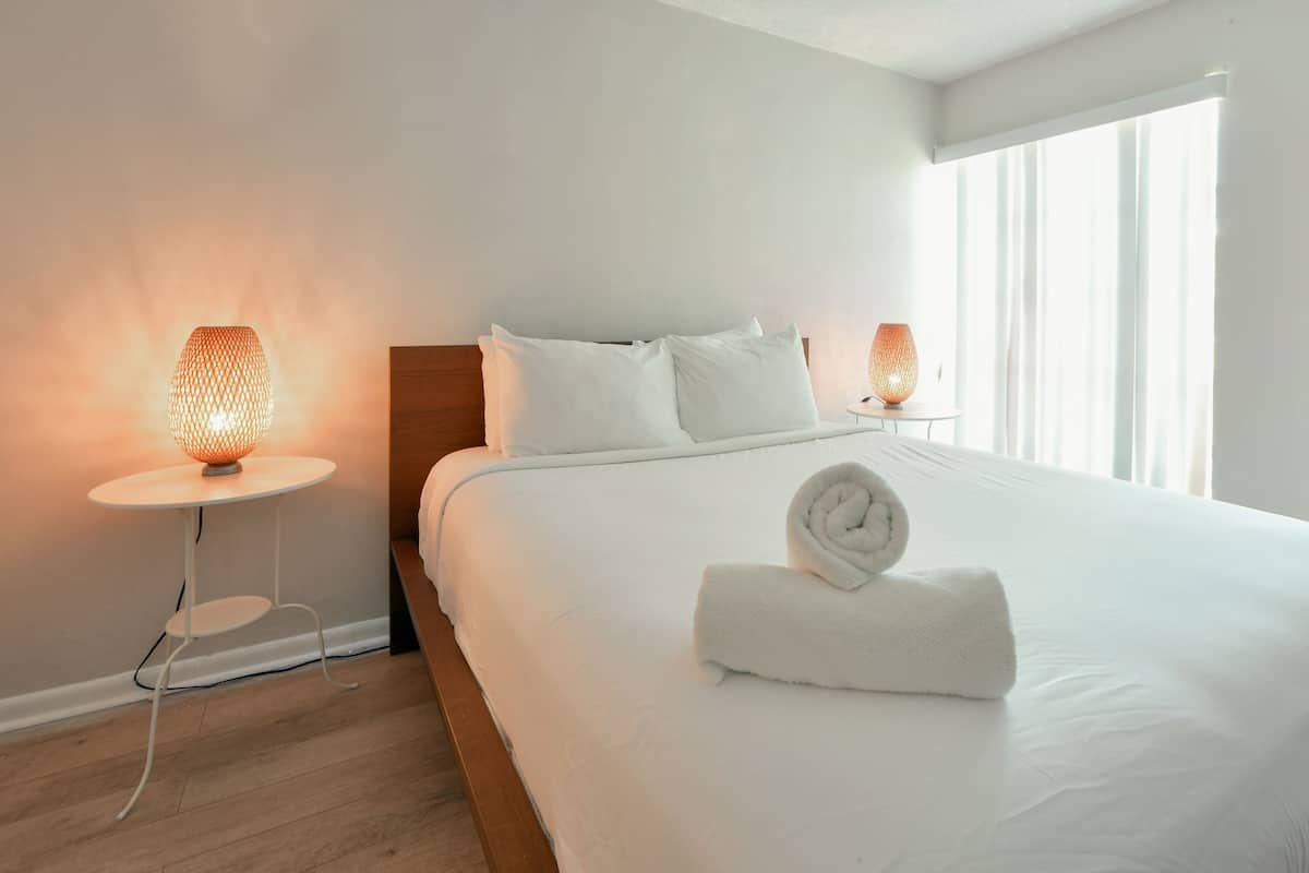 A neatly made bed with white linens and a rolled towel, two bedside lamps, and a window with sheer curtains.