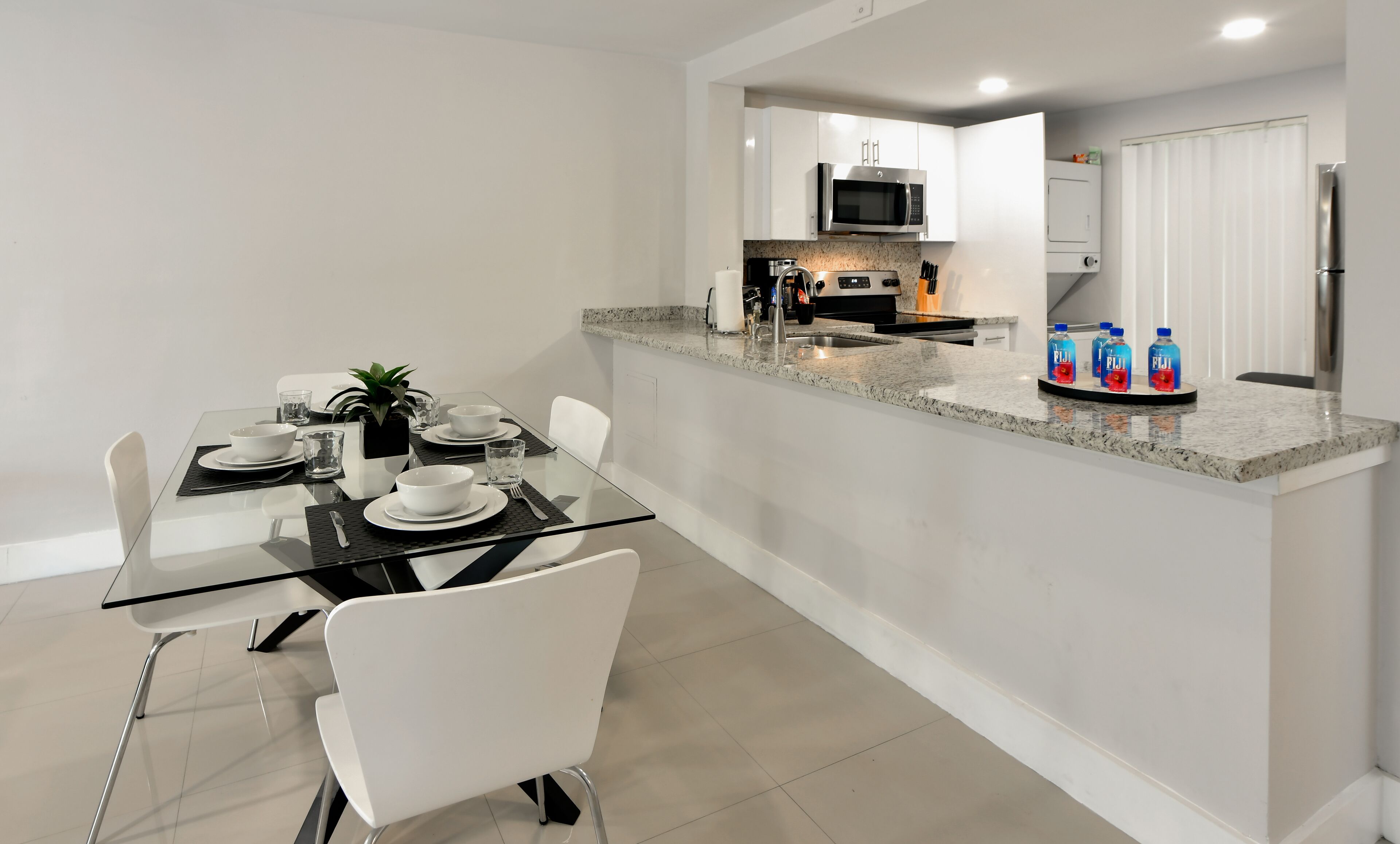 Een moderne keuken met een magnetron, kookplaat en koelkast. Er is een eethoek met een glazen tafel en witte stoelen.