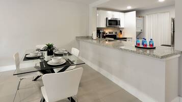 Een moderne keuken met een magnetron, kookplaat en koelkast. Er is een eethoek met een glazen tafel en witte stoelen.