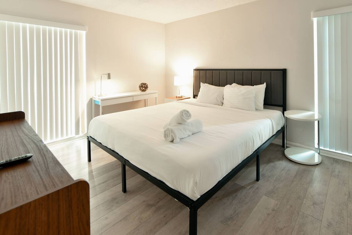 A neatly made bed with white linens and rolled towels, a bedside table with a lamp, and a round side table.