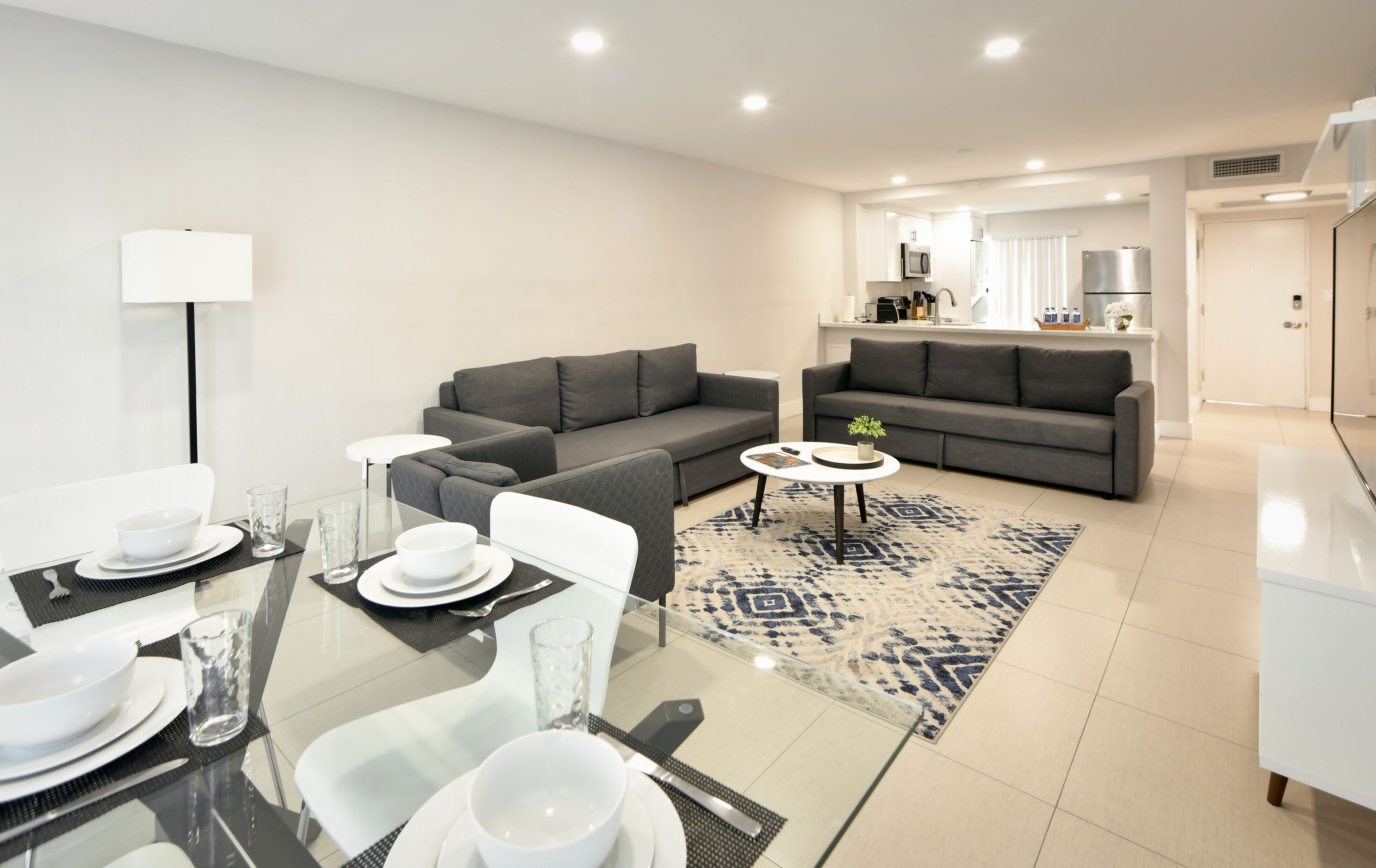 A modern living room with two grey sofas, a glass dining table set with plates and glasses, and a patterned rug.