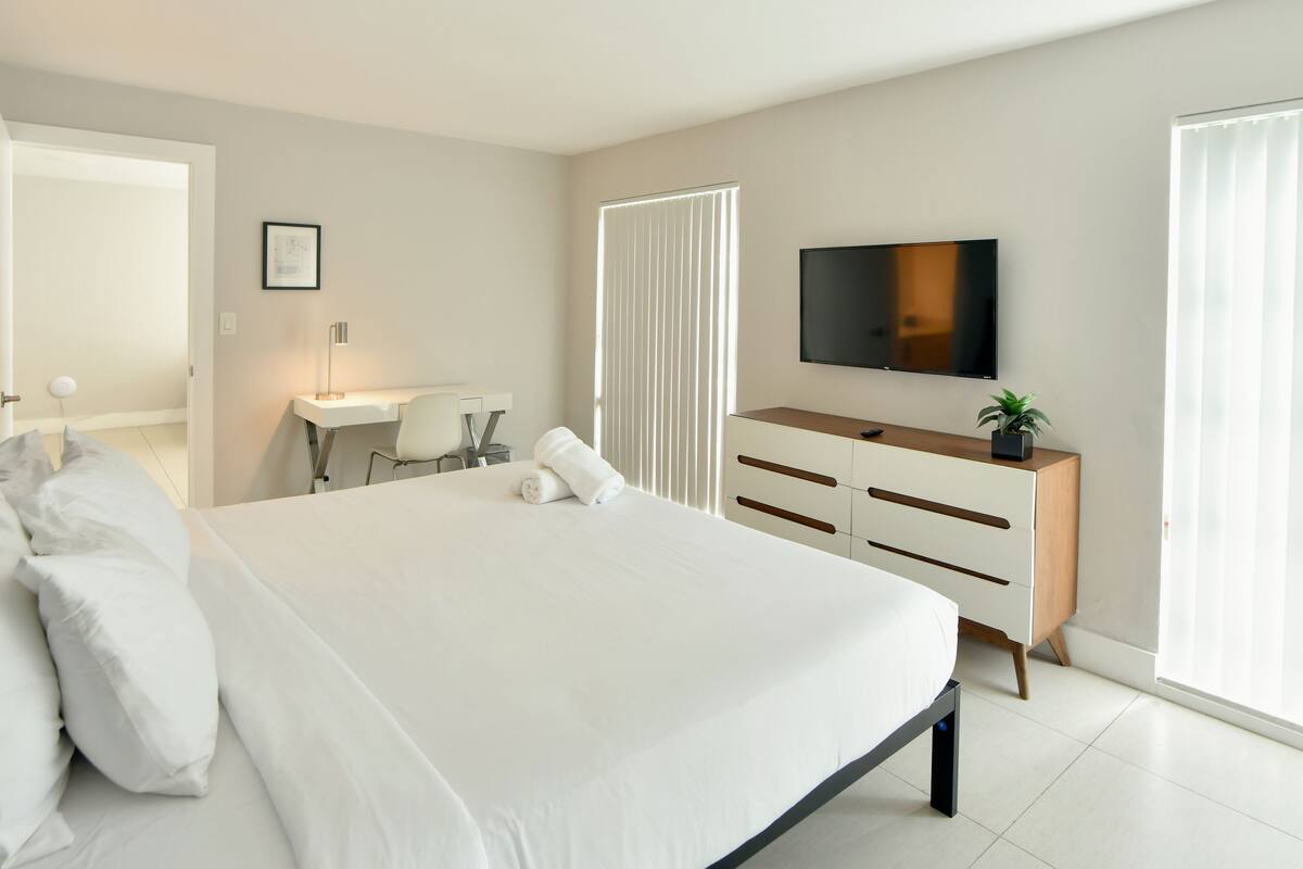 A modern hotel room with a large bed, a desk, a chair, a TV, and a window with blinds.